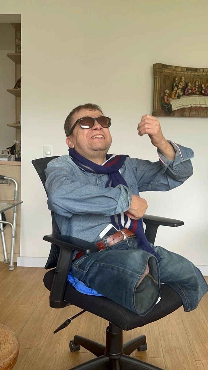 Jorge sonríe, sentado en una silla, mientras levanta una mano.